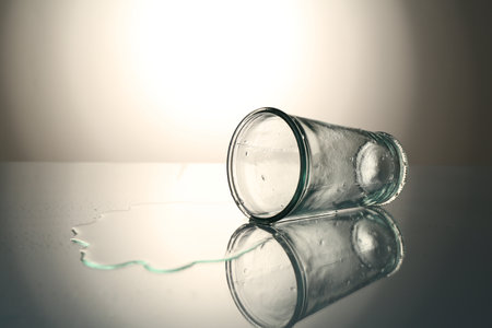 Overturned glass and spilled water on mirror table against gray gradient background, space for textの写真素材