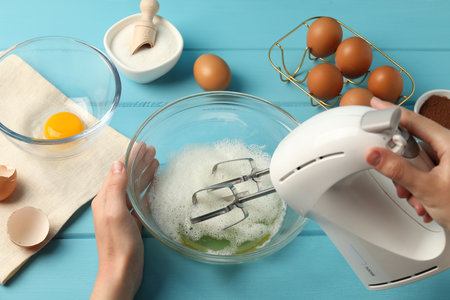 Woman whisking egg whites with hand mixer at light blue wooden table, top viewの写真素材