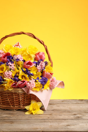 Wicker basket with beautiful flowers on wooden table against yellow backgroundの写真素材