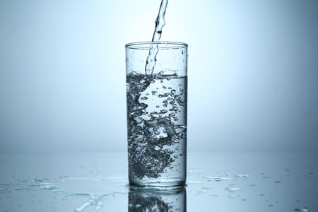Pouring water into glass at mirror table against light grey gradient background, closeup