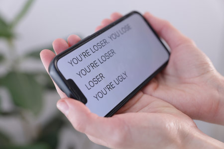 Cyber bullying. Woman holding smartphone with insults on blurred background, closeupのeditorial素材