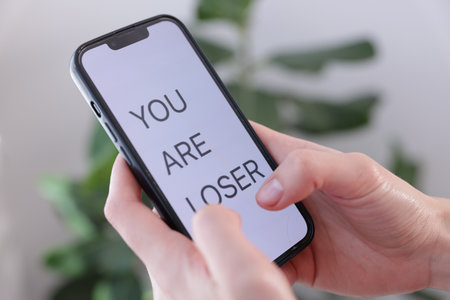Cyber bullying. Woman holding smartphone with text You Are Loser on blurred background, closeupの写真素材