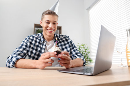 Birthday celebration. Man with cupcake having video call via laptop at table indoors, low angle viewの写真素材