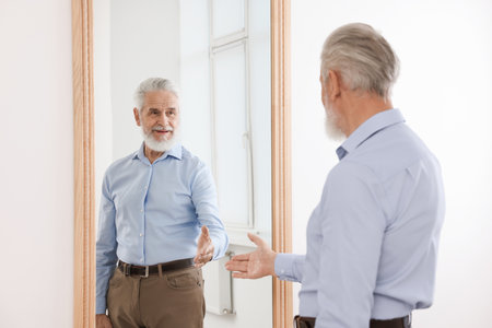 Handsome senior man looking at himself in mirror indoorsの写真素材