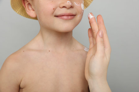 Mother applying sun protection cream onto her son's skin on light grey background, closeup