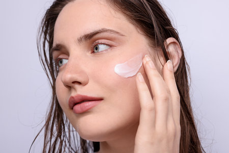 Beautiful woman applying skincare product onto her face on light grey background, closeupの写真素材