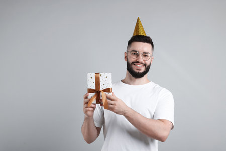 Happy Birthday. Handsome man with party hat and gift box on grey background, space for textの写真素材