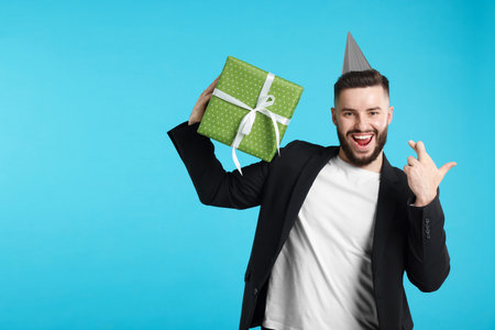 Happy Birthday. Handsome man with party hat and gift box crossing fingers while making wish on light blue background, space for textの写真素材