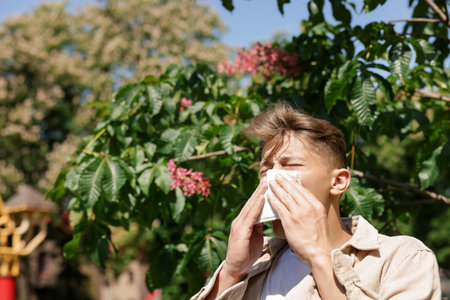 Young man with paper tissue suffering from seasonal pollen allergy near blossoming tree on spring...