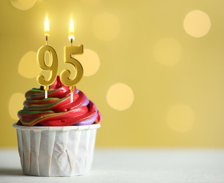 Yummy cupcake with burning number shaped candles on yellow background with blurred lights, space for text. 95th birthday or other special occasionの写真素材