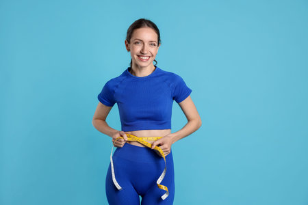 Beautiful woman measuring her belly with tape on light blue background