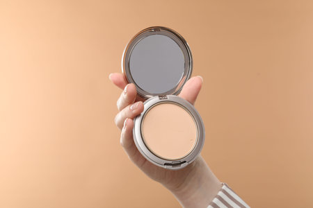 Woman holding open face powder with mirror on beige background, closeup
