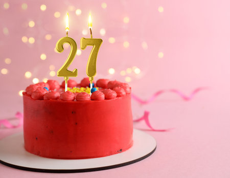 Yummy cake with burning number shaped candles on pink background with blurred lights, space for text. 27th birthday or other special occasionの写真素材