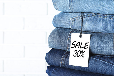Sale tag and stack of jeans against white brick wall, closeup