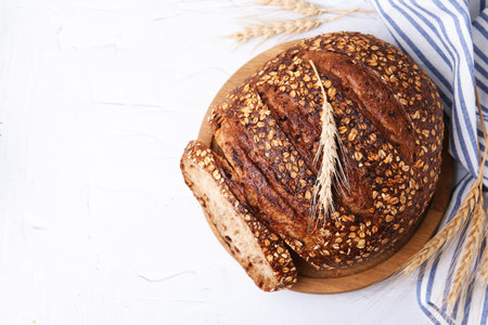 Freshly baked loaf of bread with oat flakes and spikes on light textured table, flat lay. Space for...