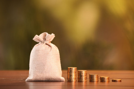 Burlap bag and stacked coins on wooden table against blurred background