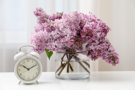 Beautiful lilac flowers in glass vase and alarm clock on table indoorsの写真素材