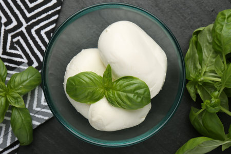 Pieces of delicious mozzarella cheese and basil in bowl on dark textured table, flat layの写真素材