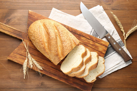 Cut bread loaf, spikelets and knife on wooden table, flat lay