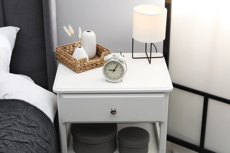 Bedside table with lamp, alarm clock and decor near bed indoors