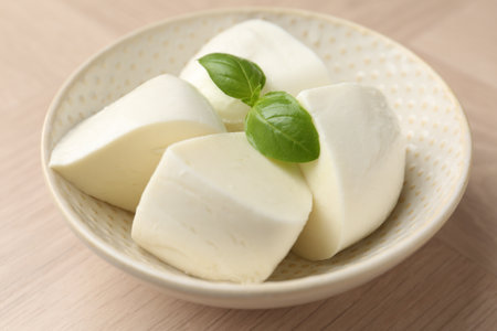 Pieces of delicious mozzarella cheese and basil in bowl on wooden table, closeupの写真素材