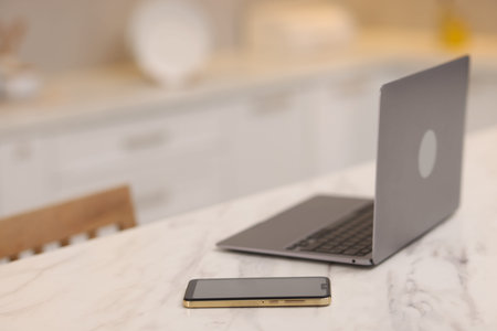 Laptop and smartphone on white marble table in kitchen, selective focus