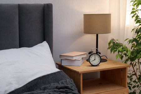 Bedside table with lamp, alarm clock and books near bed indoors. Interior design