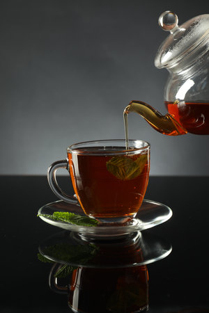 Pouring freshly brewed tea from teapot into cup at dark mirror table, closeupの写真素材