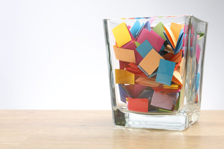 Jar with colorful pieces of paper on wooden table against light background. Space for text