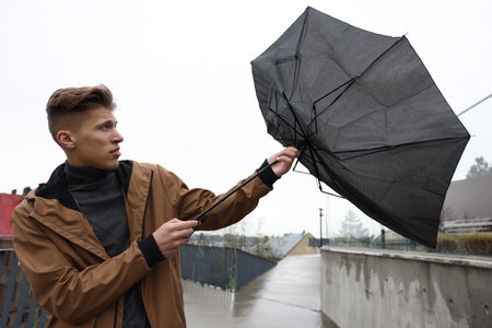 Man with broken umbrella fighting wind under rain outdoors