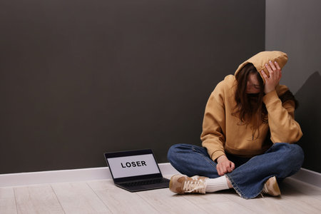 Cyber bullying. Upset teenage girl and laptop with text You Loser on floor indoors. Space for text