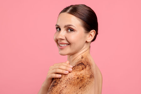 Smiling woman applying body scrub onto her shoulder against pink backgroundの写真素材