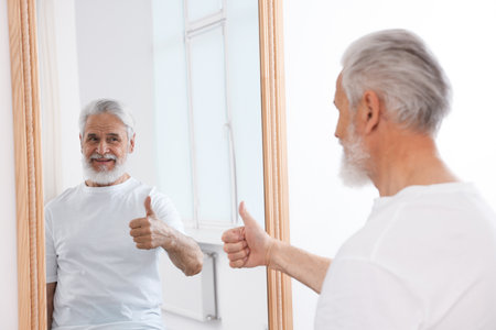 Handsome senior man showing thumbs up near mirror indoorsの写真素材