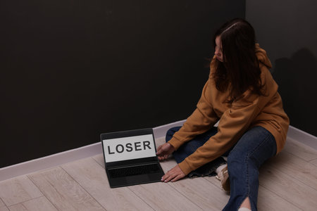 Cyber bullying. Upset teenage girl and laptop with text You Loser on floor indoors. Space for text
