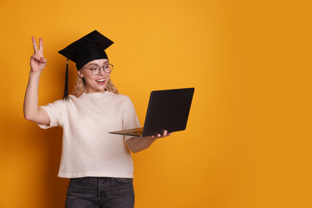 Happy student with laptop showing peace sign after graduation on orange background. Space for textの写真素材