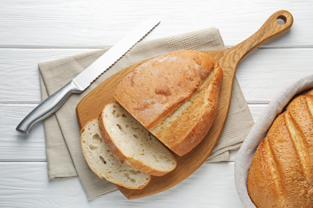 Cut bread loaf and knife on white wooden table, flat lay