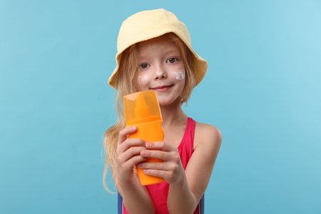 Cute little girl with bottle of sun protection product on light blue background