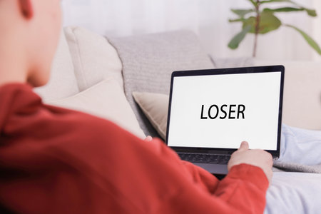 Cyber bullying. Upset teenage boy looking at laptop with text You Loser on sofa at home, closeup