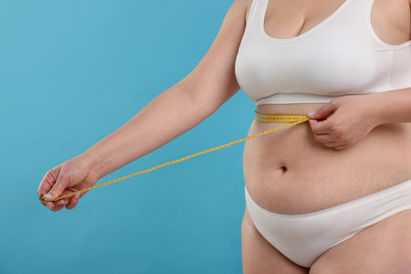 Weight loss. Plus size woman measuring waist with tape on light blue background, closeupの写真素材
