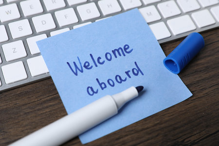 Paper note with phrase Welcome Aboard, marker and keyboard on wooden table, closeupの写真素材