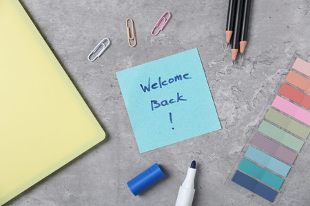 Sticky note with phrase Welcome Back and different stationery on grey table, flat lay
