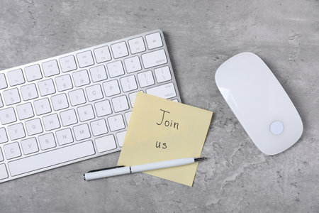 Sticky note with phrase Join Us, computer keyboard, mouse and pen on grey table, flat layの写真素材