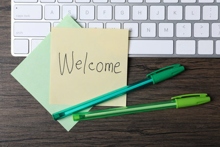 Paper note with word Welcome, pens and keyboard on wooden table, flat layの写真素材