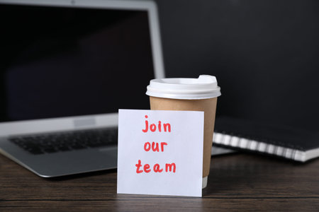 Paper note with phrase Join Our Team, cup of coffee, laptop and notebook on wooden table