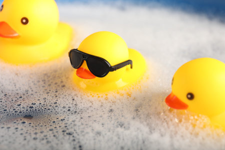 Cute rubber ducks floating in water with foam, closeup