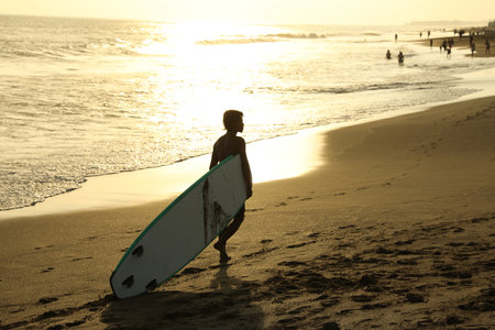 Bali, Indonesia - May 20, 2025: Man with surfboard walking along seashore at sunriseの写真素材