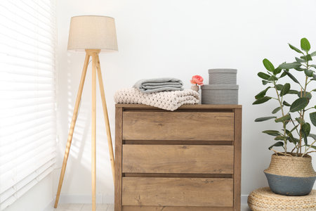 Woolen blankets on wooden chest of drawers, lamp and houseplant near white wall indoorsの写真素材