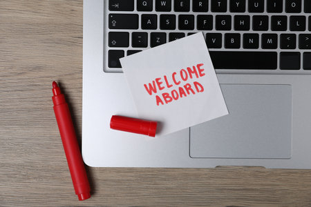 Sticky note with phrase Welcome Aboard, laptop and marker on wooden table, flat layの写真素材