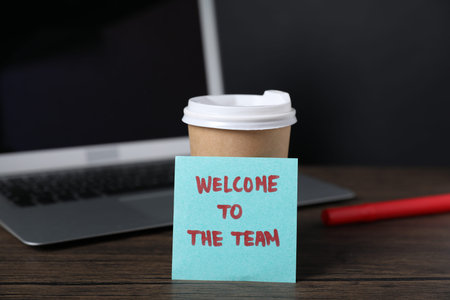 Paper note with phrase Welcome To The Team, cup of coffee, pen and laptop on wooden tableの写真素材