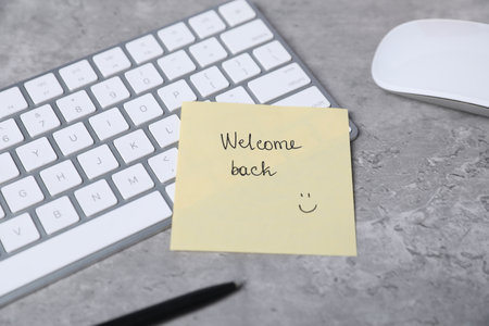 Sticky note with phrase Welcome Back, computer keyboard, mouse and pen on grey table, closeupの写真素材